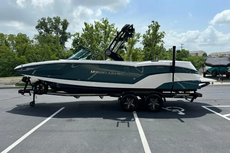Slide: The Image of 2025 MasterCraft NXT24 boat on trailer in parking lot, surrounded by trees and cloudy sky. - 6