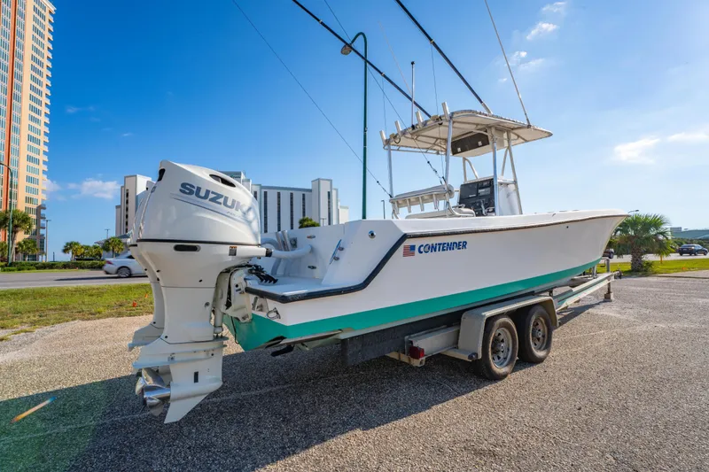 Slide: The Image of 1997 Contender 27 boat on trailer with Suzuki outboard, parked near buildings under blue sky. - 3