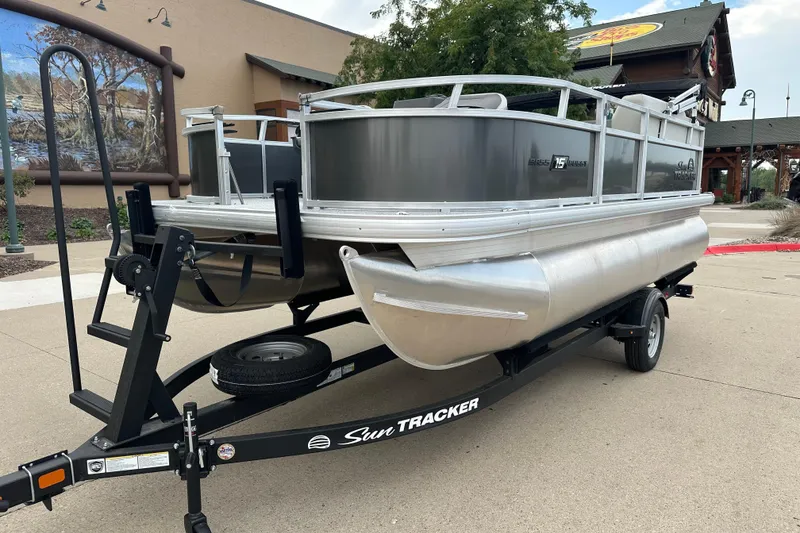 The Image of 2026 Sun Tracker Bass Buggy 16 XL Select pontoon boat on trailer, parked outdoors. - 0