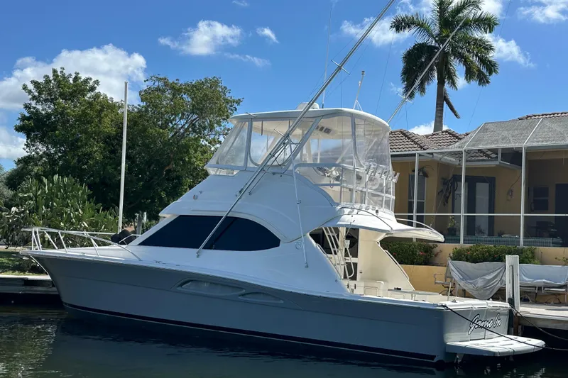 Slide: The Image of 2002 Riviera 40 Flybridge yacht docked near a waterfront home, under a clear blue sky. - 1