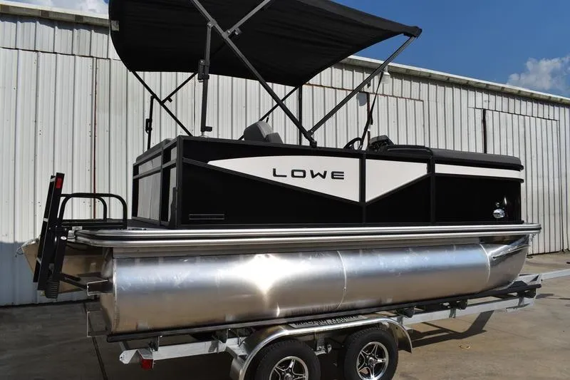 The Image of 2026 Lowe LS 170 pontoon boat with canopy on trailer, parked by a warehouse. - 0