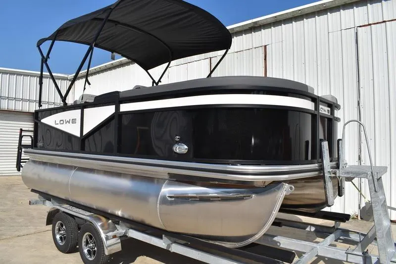 Slide: The Image of 2026 Lowe LS 170 pontoon boat with black canopy on trailer, parked by a white building. - 2
