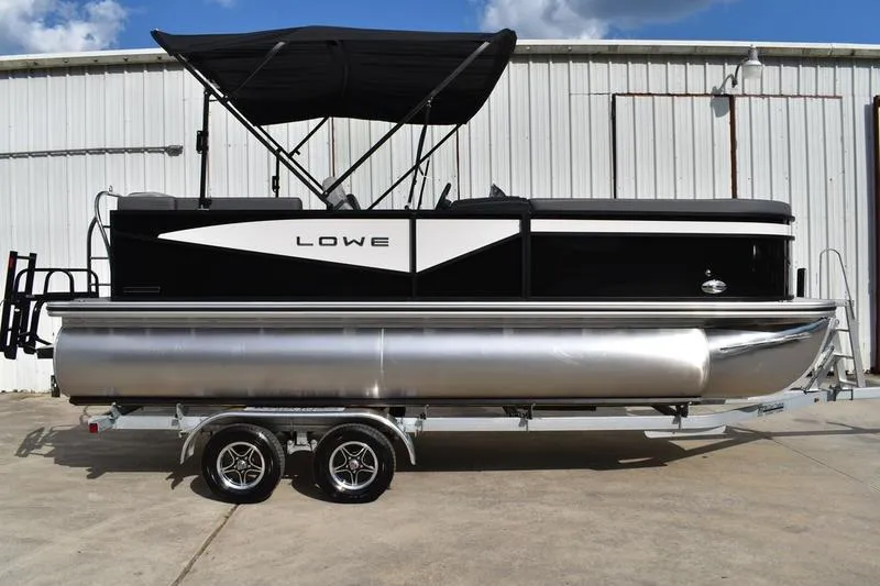 Slide: The Image of 2026 Lowe LS 210 WT pontoon boat with canopy on trailer, parked by a warehouse. - 2