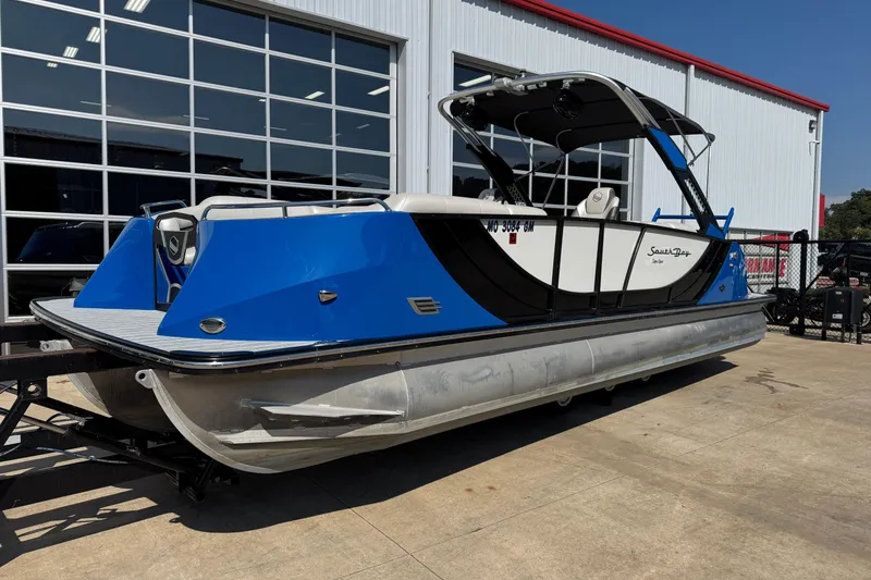 Slide: The Image of 2022 South Bay 25 Sport CR9-DC pontoon boat with blue accents, parked outside a building. - 2