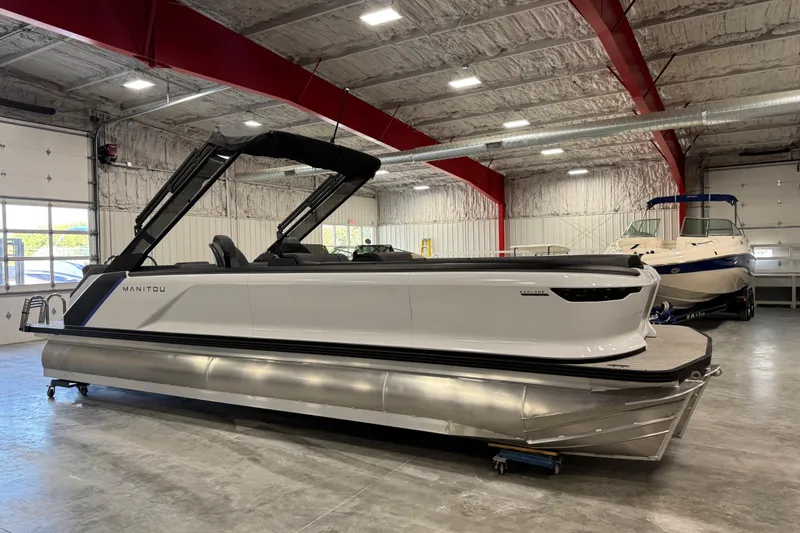 Slide: The Image of 2025 Manitou 26 Explore pontoon boat in a spacious indoor showroom. - 1