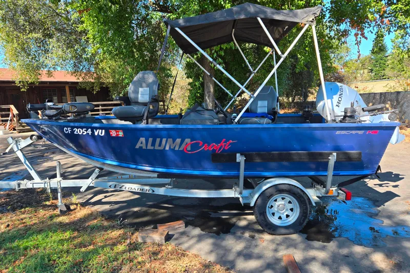 Slide: The Image of 2022 Alumacraft Escape 145 CS boat on trailer, parked outdoors under trees. - 2