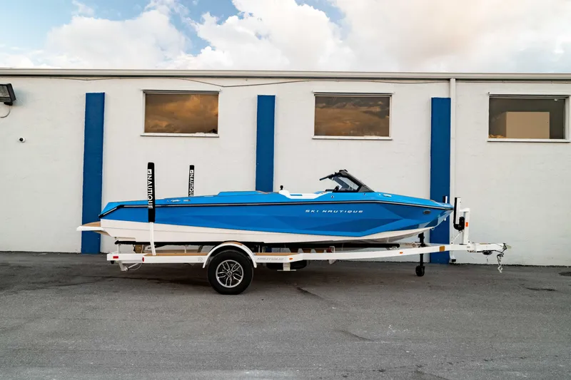 The Image of 2026 Nautique Ski Nautique boat in blue on trailer, parked by a building. - 0
