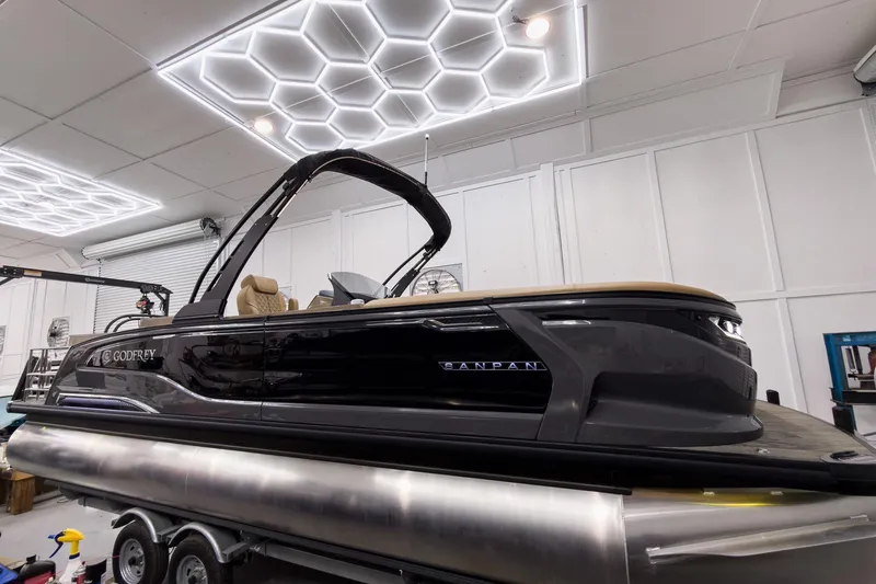 Slide: The Image of 2026 Godfrey Sanpan 25' Tritoon boat in a modern showroom with hexagonal ceiling lights. - 11