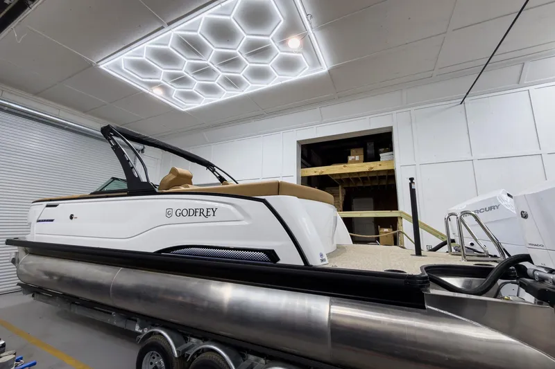 Slide: The Image of 2026 Godfrey Sanpan 27' Tritoon boat in a modern showroom with hexagonal ceiling lights. - 11