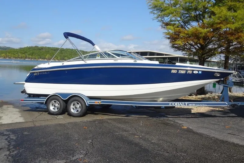 Slide: The Image of 2003 Cobalt 24SD boat on trailer by a lake, blue and white design. - 1