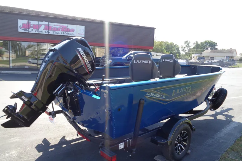 Slide: The Image of 2026 Lund 1650 Rebel Tiller boat with Mercury engine, parked outside a marine store. - 3