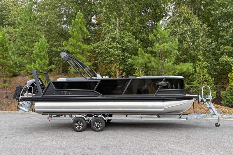 The Image of 2026 Alera M23Q pontoon boat on trailer, surrounded by lush green trees. - 1