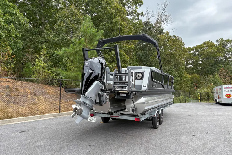 Slide: The Image of 2026 Alera M23Q pontoon boat on trailer, parked near wooded area. - 2