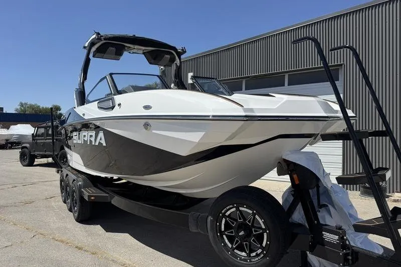 Slide: The Image of 2025 Supra SE 575 boat on trailer, parked outside industrial building. - 2