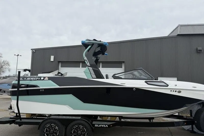 The Image of 2025 Supra SL 575 boat on trailer, sleek design, black and teal accents, parked near industrial building. - 0