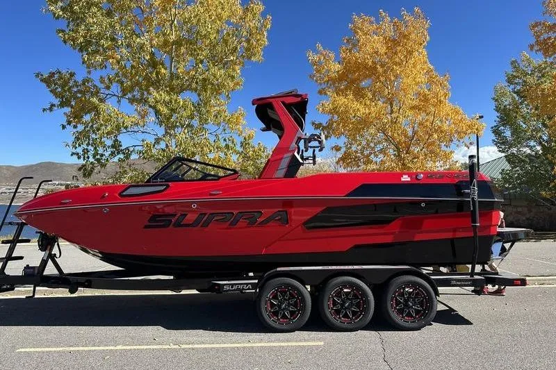 The Image of 2024 Supra SE 575 boat in vibrant red on a trailer, surrounded by autumn trees. - 0