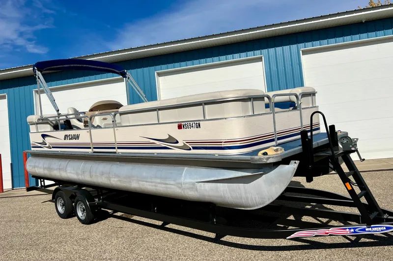 The Image of 2003 Sylvan 824 Mirage Cruise pontoon boat on trailer, parked near blue building. - 0