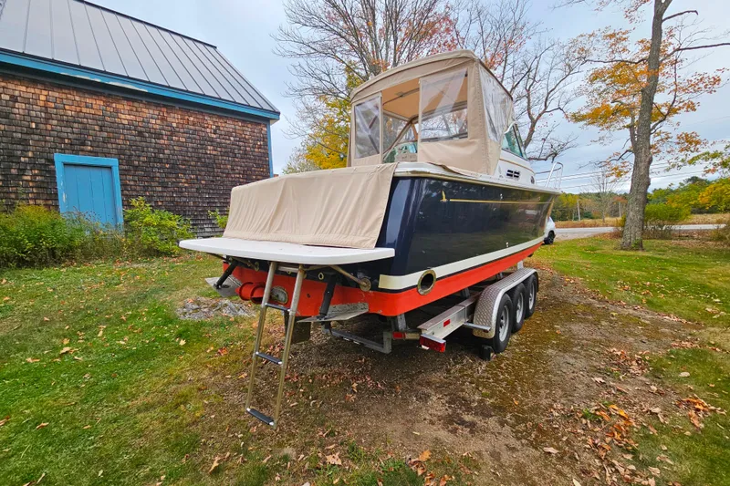 Slide: The Image of 2006 Back Cove 26 boat on trailer, parked on grassy area near wooden building. - 30