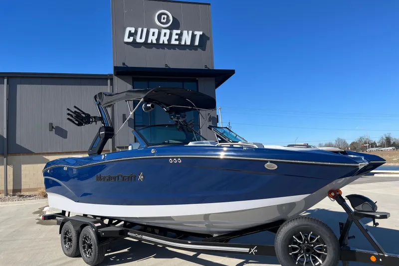 The Image of 2025 MasterCraft X24 boat on trailer, parked outside showroom under clear blue sky. - 0