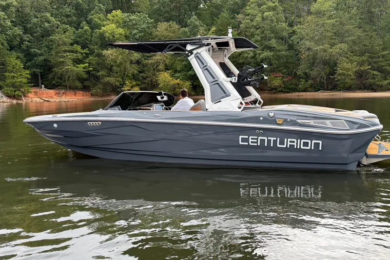 Slide: The Image of 2026 Centurion Nv233 boat on a calm lake, surrounded by lush green trees. - 2
