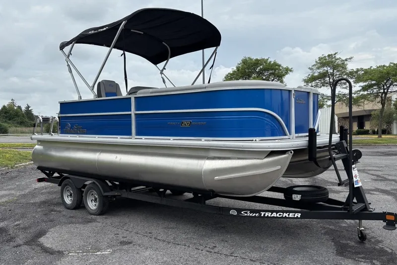 Slide: The Image of 2026 Sun Tracker Party Barge 20 DLX pontoon boat on trailer, blue exterior, parked outdoors. - 2