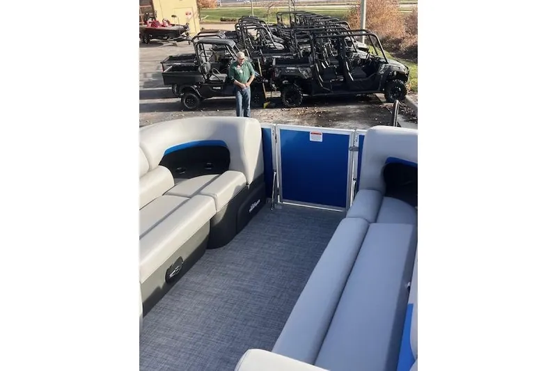 Slide: The Image of 2026 Sun Tracker Party Barge 20 DLX interior with gray seating, parked utility vehicles in background. - 72