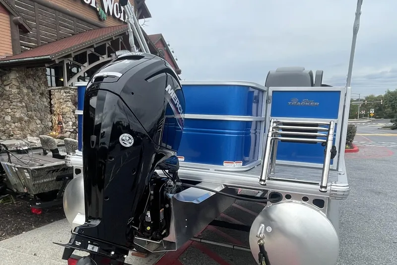 Slide: The Image of 2026 Sun Tracker Party Barge 18 DLX with Mercury outboard motor, parked outside a rustic building. - 2