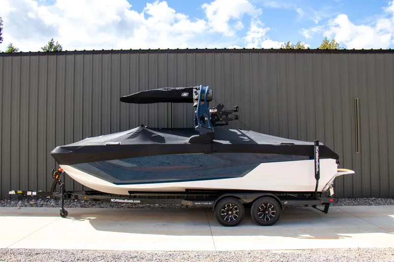 Slide: The Image of 2026 Nautique Super Air Nautique GS24 boat on trailer, covered, against industrial building. - 6