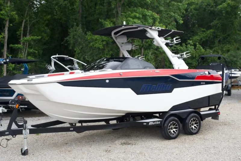 The Image of 2021 Malibu M220 boat on trailer, parked outdoors with lush green trees in background. - 1