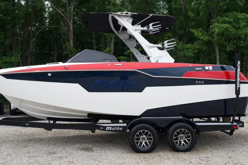 Slide: The Image of 2021 Malibu M220 boat on trailer, sleek design, red and blue accents, forest background. - 2