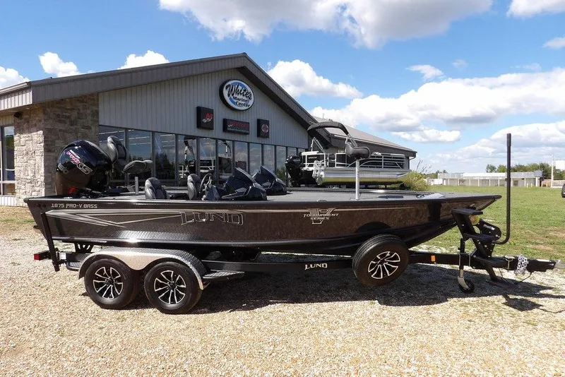 Slide: The Image of 2026 Lund 1875 Pro-V Bass XS boat parked outside a marine center under a blue sky. - 4