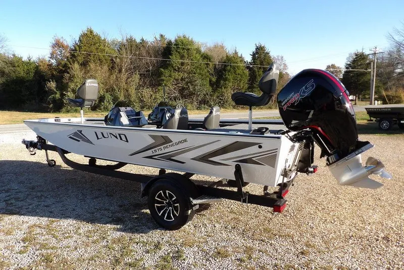 Slide: The Image of 2026 Lund 1975 Renegade boat with motor, parked on gravel, surrounded by trees. - 9