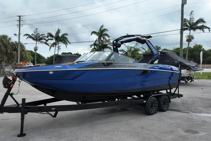 Slide: The Image of 2025 Centurion Fe23 boat in blue on trailer, parked outdoors with palm trees. - 4