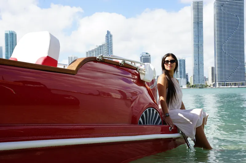 Slide: The Image of Woman on a red Car Off-shore Fiat 500 Classic boat, 2025, with city skyline backdrop. - 13