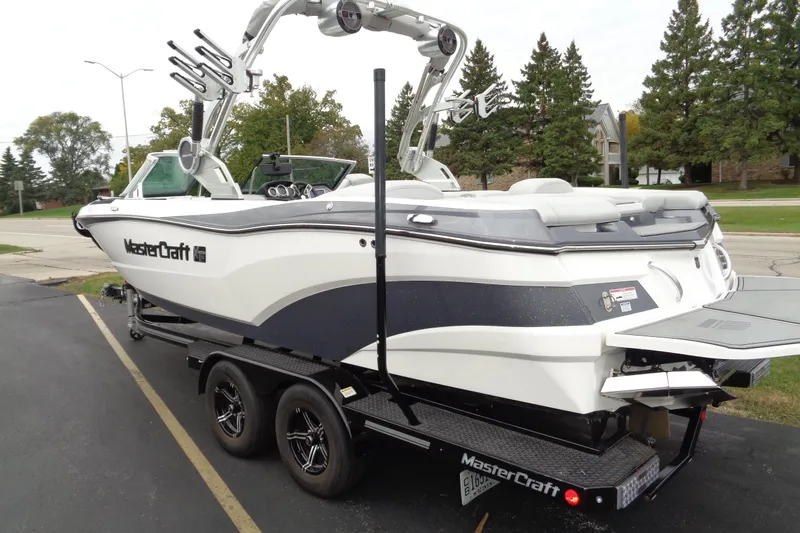 Slide: The Image of 2020 MasterCraft XT23 boat on trailer, parked outdoors with trees in background. - 47