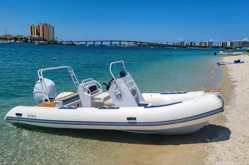 The Image of 2026 East Marine 560 boat on sandy beach with clear blue water and cityscape background. - 0