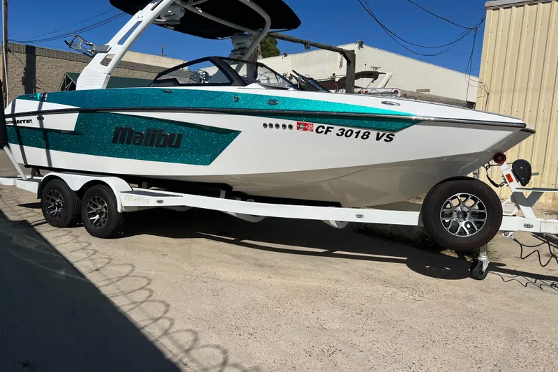 Slide: The Image of 2020 Malibu Wakesetter 23 MXZ boat on trailer, teal and white design, parked outdoors. - 2