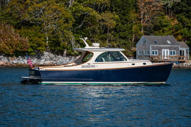 Slide: The Image of 2013 Hinckley Talaria 34 boat cruising near a lakeside house with trees in the background. - 2