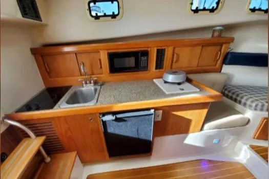 Slide: The Image of 1999 Mainship Pilot 30 interior with kitchenette, featuring wooden cabinetry, sink, microwave, and seating area. - 24