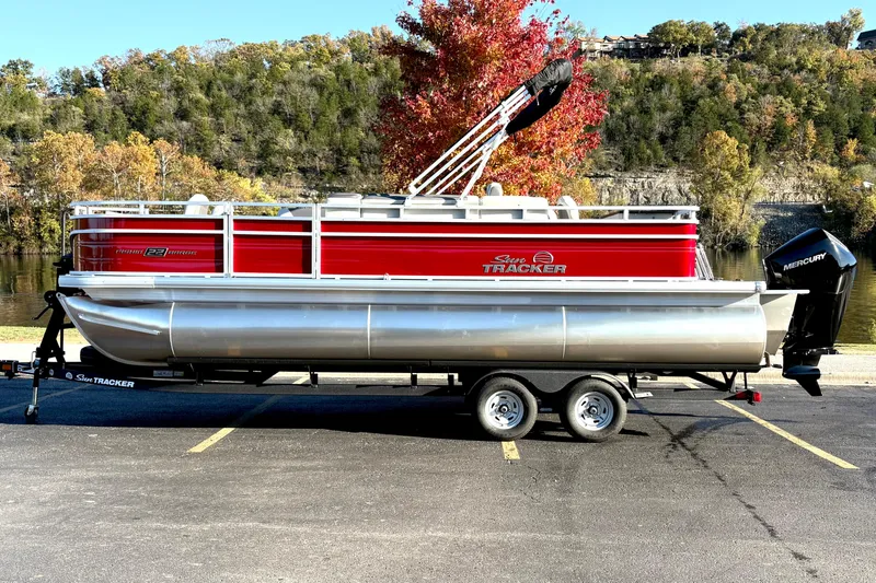 Slide: The Image of 2026 Sun Tracker Fishin' Barge 22 XP3 pontoon boat on trailer, parked by scenic lake. - 118