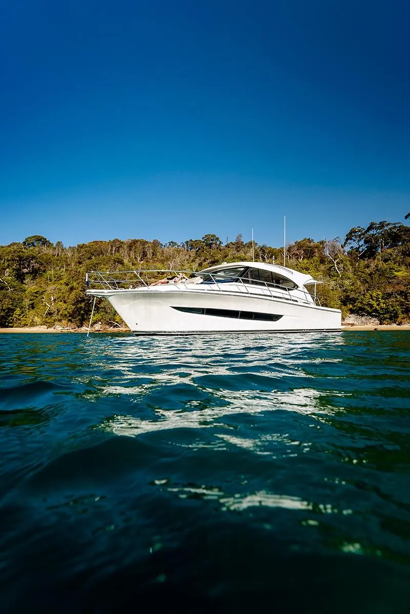 Slide: The Image of 2024 Riviera 395 SUV yacht on calm water with forested shoreline. - 2