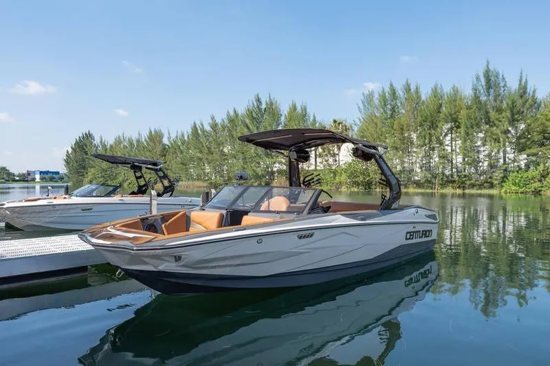 The Image of 2025 Centurion Nv233 boat docked on a serene lake with lush trees in the background. - 7