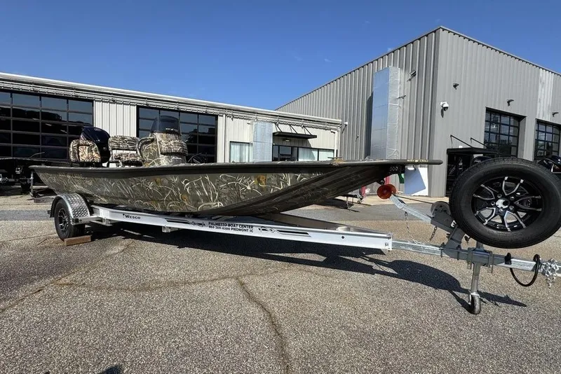 The Image of 2026 War Eagle 961 Blackhawk boat on trailer, camo design, parked outside industrial building. - 1