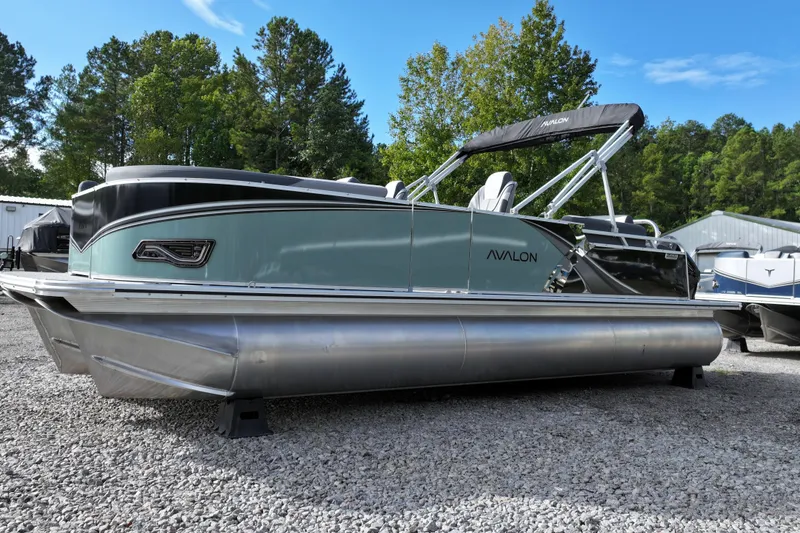 Slide: The Image of 2026 Avalon LSZ Quad Lounger Shift pontoon boat on gravel lot, surrounded by trees. - 2