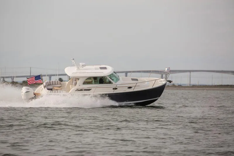 Slide: The Image of 2026 Back Cove 340 boat cruising on water with American flag, bridge in background. - 2