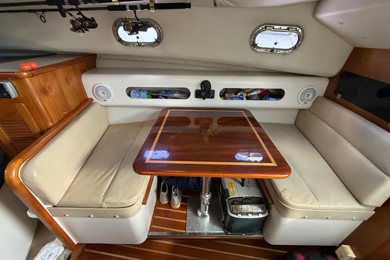 Slide: The Image of 2004 Pursuit 3100 Offshore cabin with wooden table, beige seating, and fishing gear storage. - 20