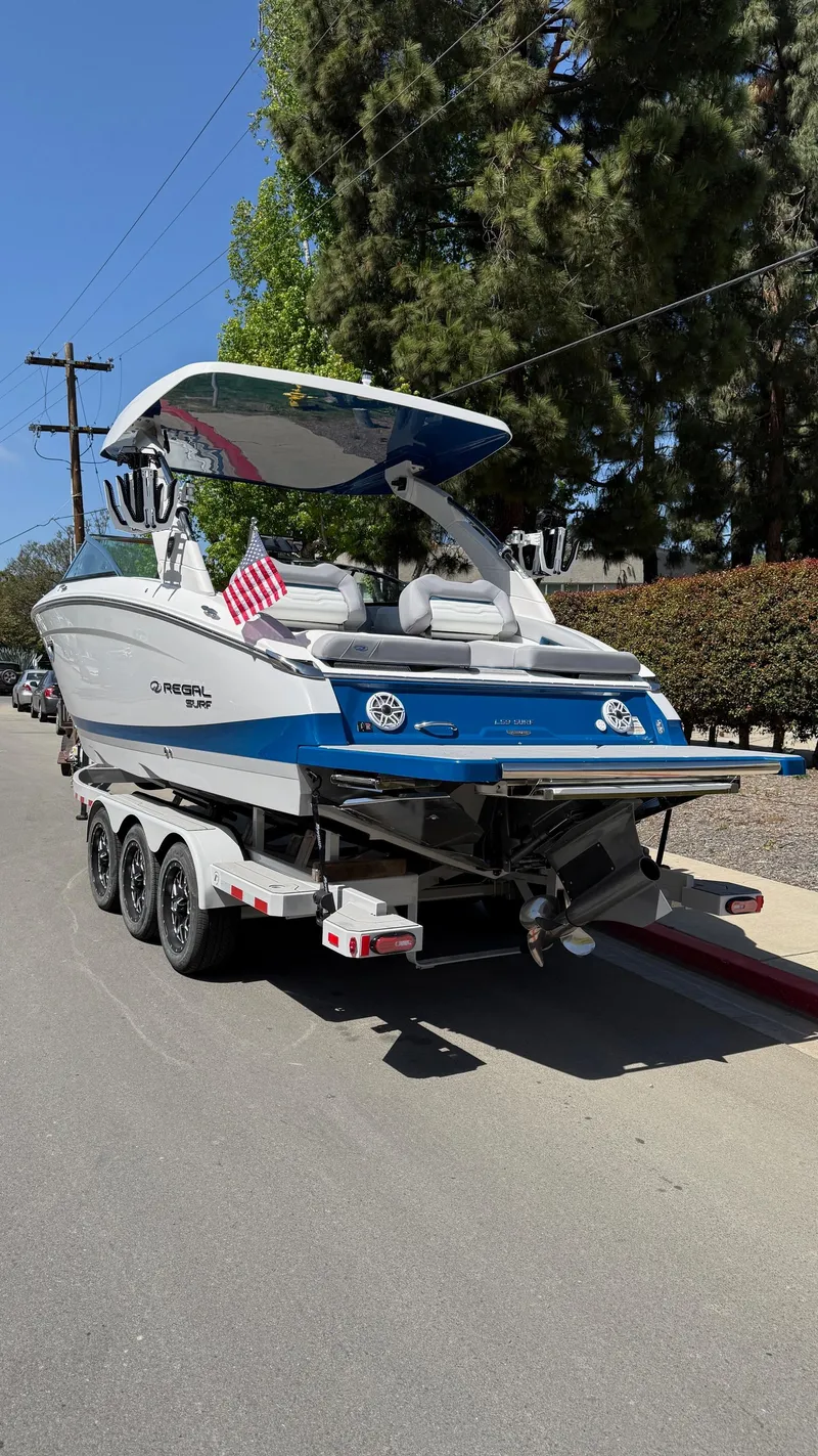 Slide: The Image of 2025 Regal LS9 Surf boat on trailer, parked on street, with trees in background. - 21
