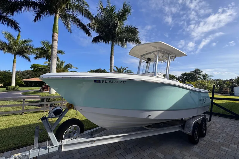 The Image of 2022 Robalo R230 Center Console boat on trailer, surrounded by palm trees and blue sky. - 0
