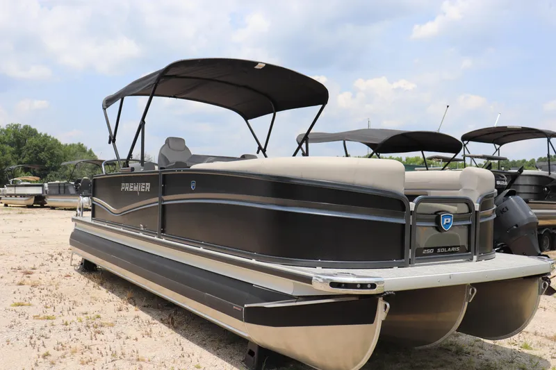 Slide: The Image of 2024 Premier 250 Solaris Rev pontoon boat with canopy, parked on sandy terrain. - 0