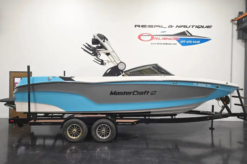 The Image of 2023 MasterCraft XT24 boat on trailer in showroom, blue and white design. - 1